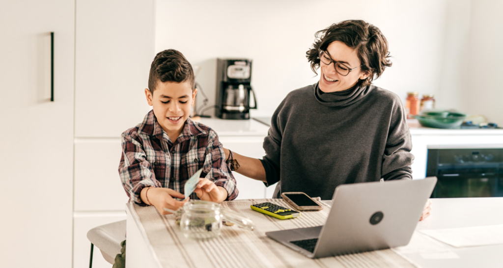 Mother and young son at kitchen table learning about savings and budgeting together. The son is holding money, ready to place it in a glass jar while the mother smiles encouragingly. A laptop, calculator, and smartphone sit on the table, emphasizing a practical lesson in financial literacy.